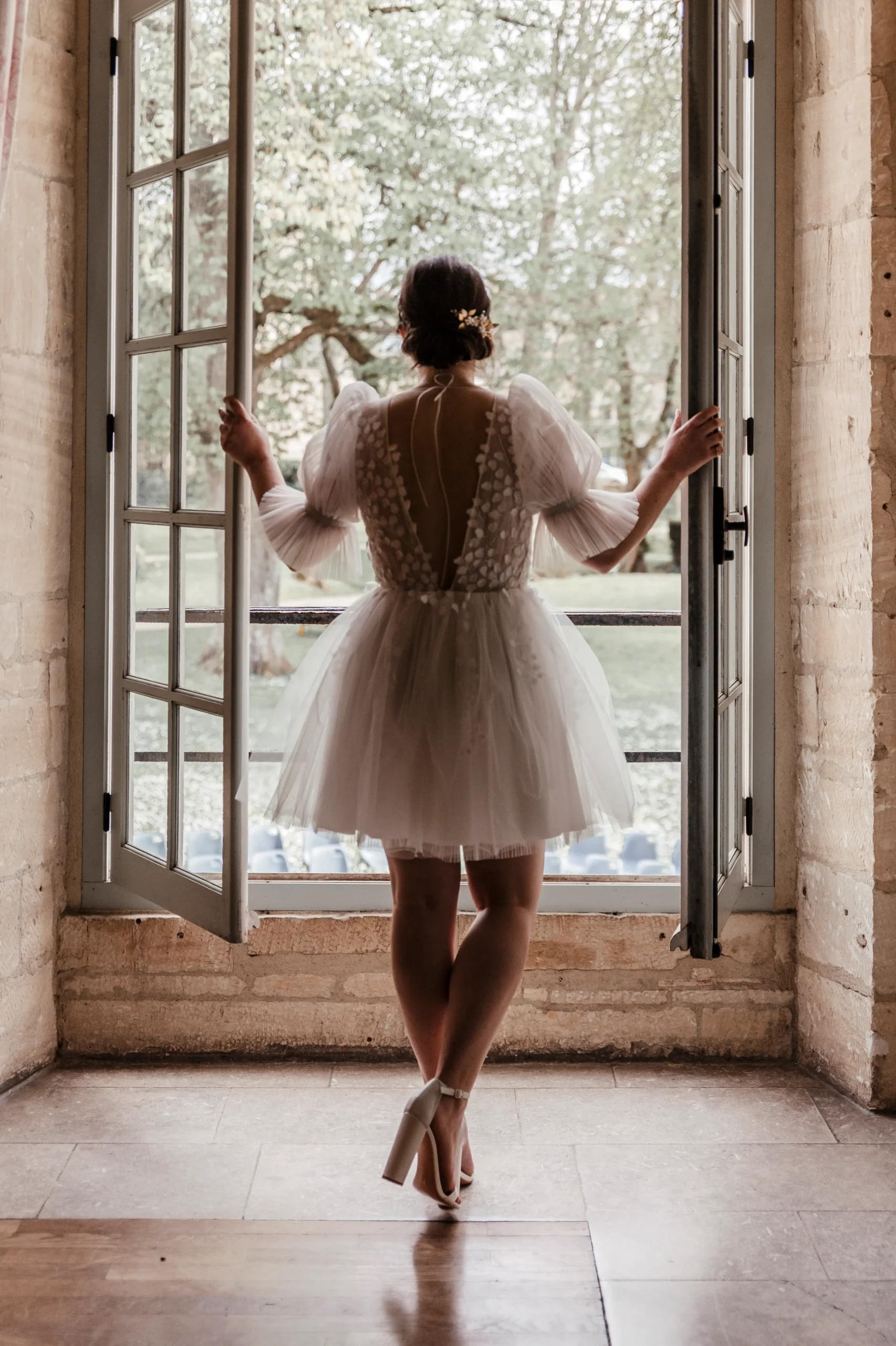 jeune mariée en robe courte et talon haut de dos devant une fenêtre ouverte au château médiéval de Creully en Normandie